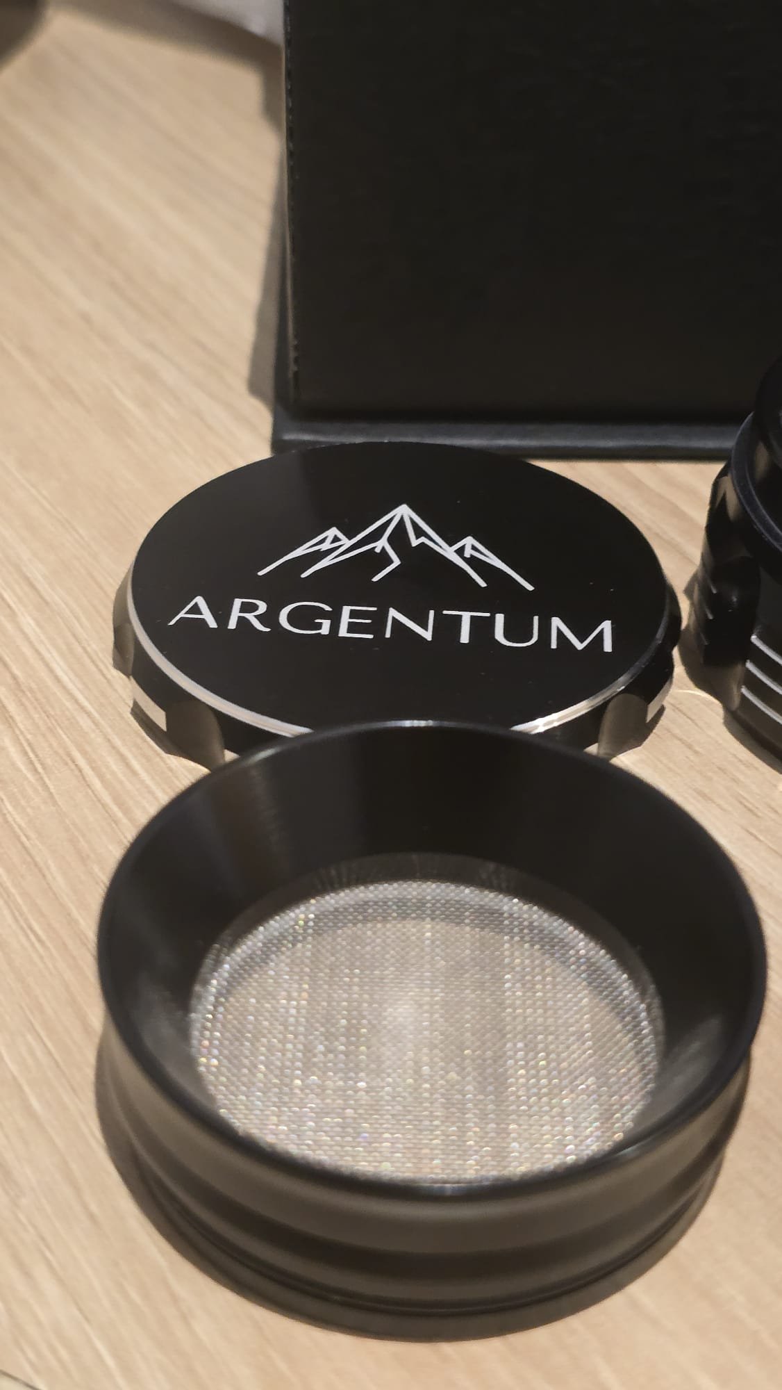Close-up of the ARGENTUM grinder's removable mesh screen chamber next to the engraved mountain logo lid
